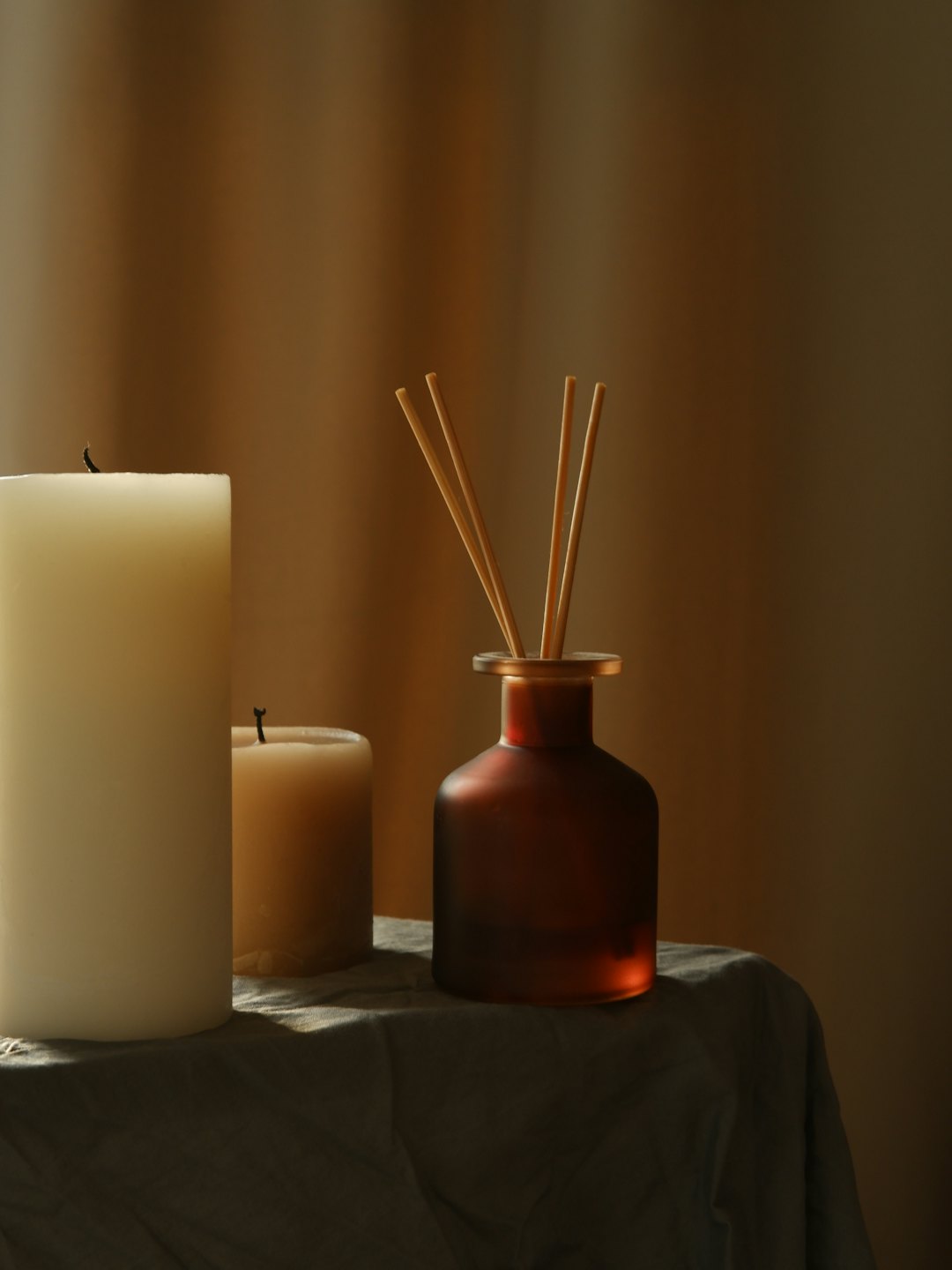 Pillar candles and reed diffuser on a draped surface.